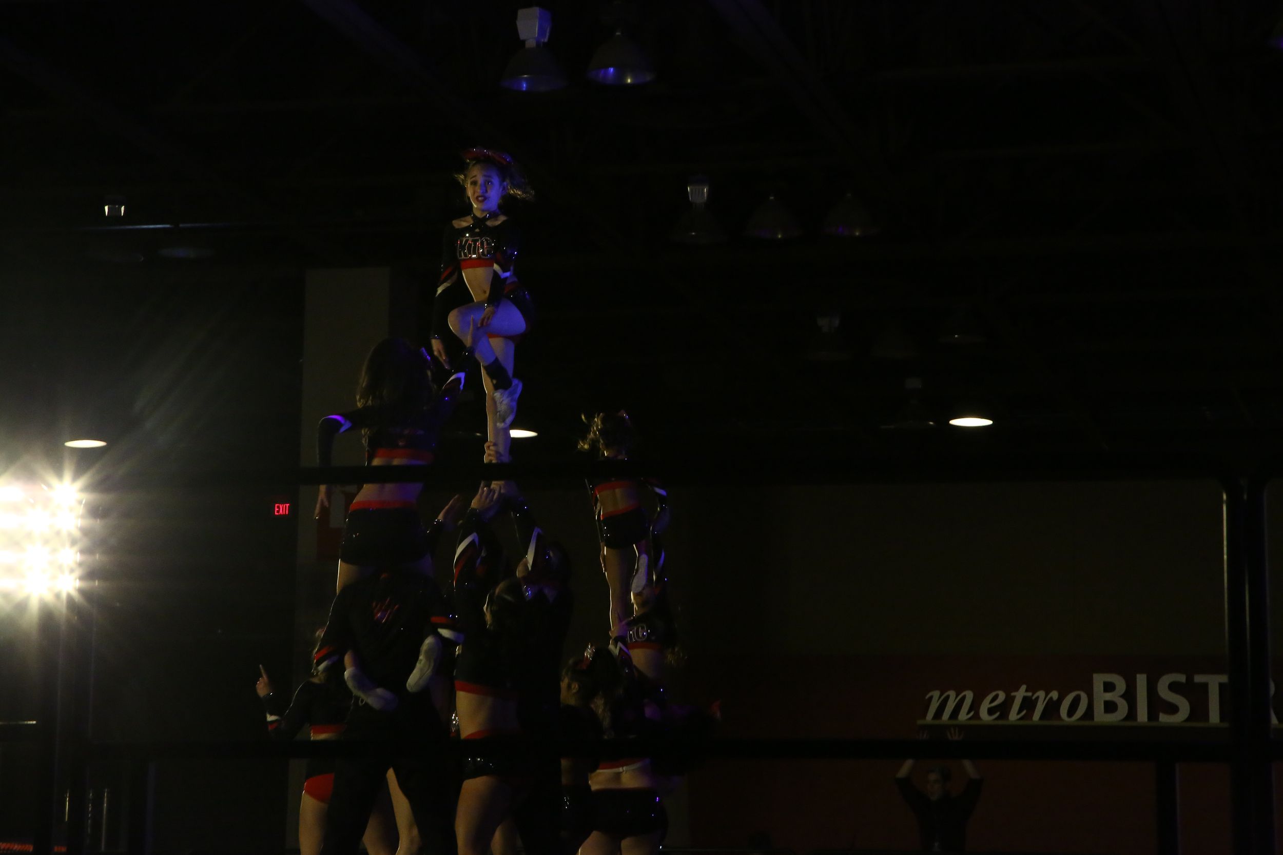 Cheerleaders form a tall pyramid stunt under dim arena lighting, with a metroBIST sign visible on the wall behind them.