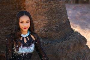 Smiling young cheerleader with long dark hair and red lipstick poses in a sparkly black uniform beside a large palm tree trunk.
