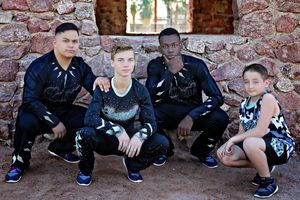 Four male cheer athletes in black team uniforms pose together in front of a rustic stone wall, with three crouching and one kneeling.