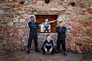Male cheer athletes in black uniforms pose at a stone wall while a young boy in a patterned outfit sits in the window opening.