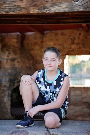 Smiling boy in a camouflage tank top and black shorts sits on a stone ledge inside a rustic stone and timber shelter.