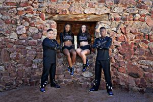 Four cheer team members in black uniforms and sneakers pose together in front of a red stone wall with a wooden beam.