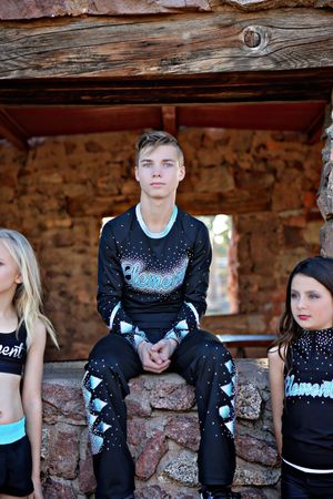 Teen boy in a black cheer uniform with blue accents sits on a stone ledge with two young cheerleaders beside him.