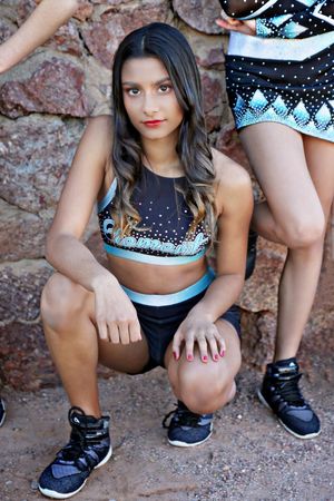 Young cheerleader in a black and teal sparkle uniform crouches on dirt in front of a red stone wall with teammates behind.