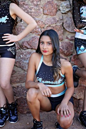 Cheerleader with long dark hair and red lipstick crouches in a black and teal crop top beside teammates by a stone wall.