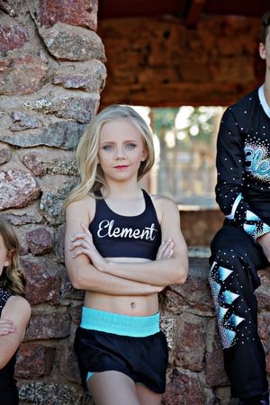 Young blonde cheerleader in a black Element crop top and teal-trimmed skirt stands with arms crossed beside a stone wall.