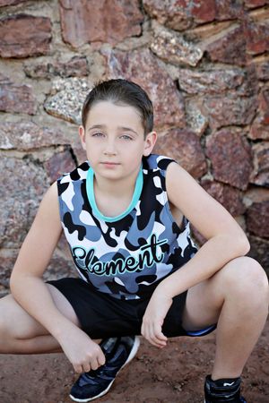 Boy in a camouflage Element tank top and black shorts crouches on red dirt in front of a rustic stone wall.