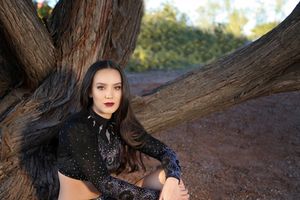 A young woman with long dark hair and red lipstick sits against a large tree trunk in a black lace cheer top.