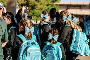 Young girls in matching teal bows and teal backpacks walk together outdoors in black team jackets on a sunny day.