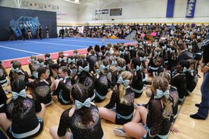 A large cheerleading team in black uniforms with teal bows sits on a gym floor watching a competition.