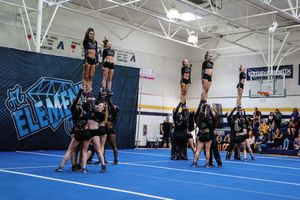 Cheerleaders in black uniforms perform a stunt formation with flyers lifted high on a blue gym mat in front of an Element banner.