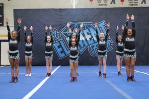 A cheer team in black and teal uniforms strikes a synchronized high-V arm pose on a blue mat in front of an Elements banner.