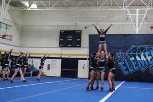 A cheerleader stands arms extended in an elevated stunt held by teammates while other athletes watch in a school gymnasium.