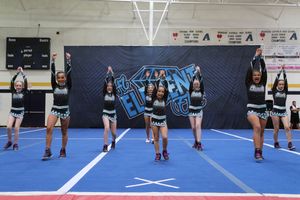 Cheer team members in black and silver uniforms stand in formation with raised arms on a blue competition floor in a gym.