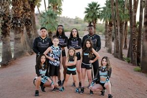 A cheer team of teens and children in black uniforms poses on a dirt path lined with palm trees in the desert.