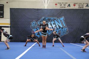 Cheer team members perform a stunt on a blue mat in a gym, with an Element banner and championship signs in the background.