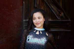 Smiling young cheerleader in a black sparkly Element uniform poses in front of a rustic weathered wooden barn door.