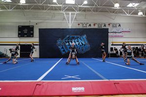 An all-star cheer team performs a routine on a blue mat with athletes spread across the floor and a red-padded edge in front.