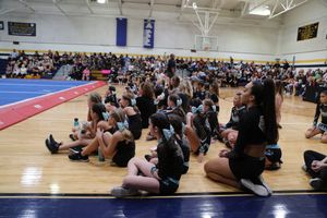 Young cheerleaders in black uniforms sit on a gym floor watching a competition while spectators fill the bleachers behind them.