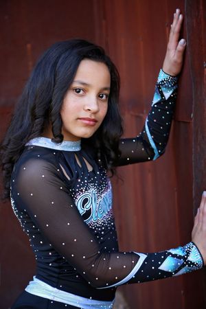 Young cheerleader in a black rhinestone uniform with blue accents leans against a rusty metal wall with her hands raised.