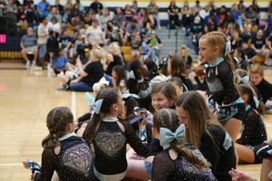 Cheerleaders in black sparkly uniforms with teal bows gather together in a gym, with a crowd of spectators in the bleachers behind.