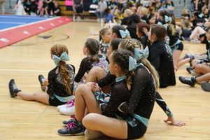 Young cheer athletes in black and teal uniforms with hair bows sit on a wood gym floor, watching a cheerleading event.