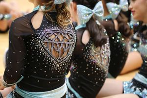 Close-up of a black cheer uniform back with a sparkling rhinestone diamond design and glittery teal hair bows on teammates.