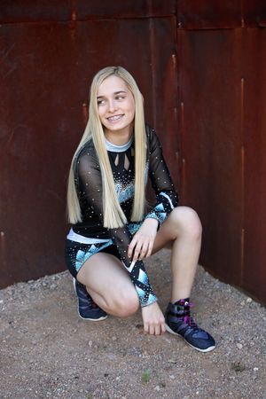 Blonde cheerleader in a black and teal uniform and sneakers crouches on gravel and smiles beside a rusty metal wall.