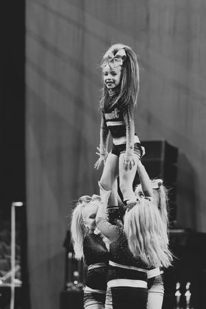 Black and white photo of a young cheerleader with a big hair bow smiling while lifted high in a stunt by her teammates.