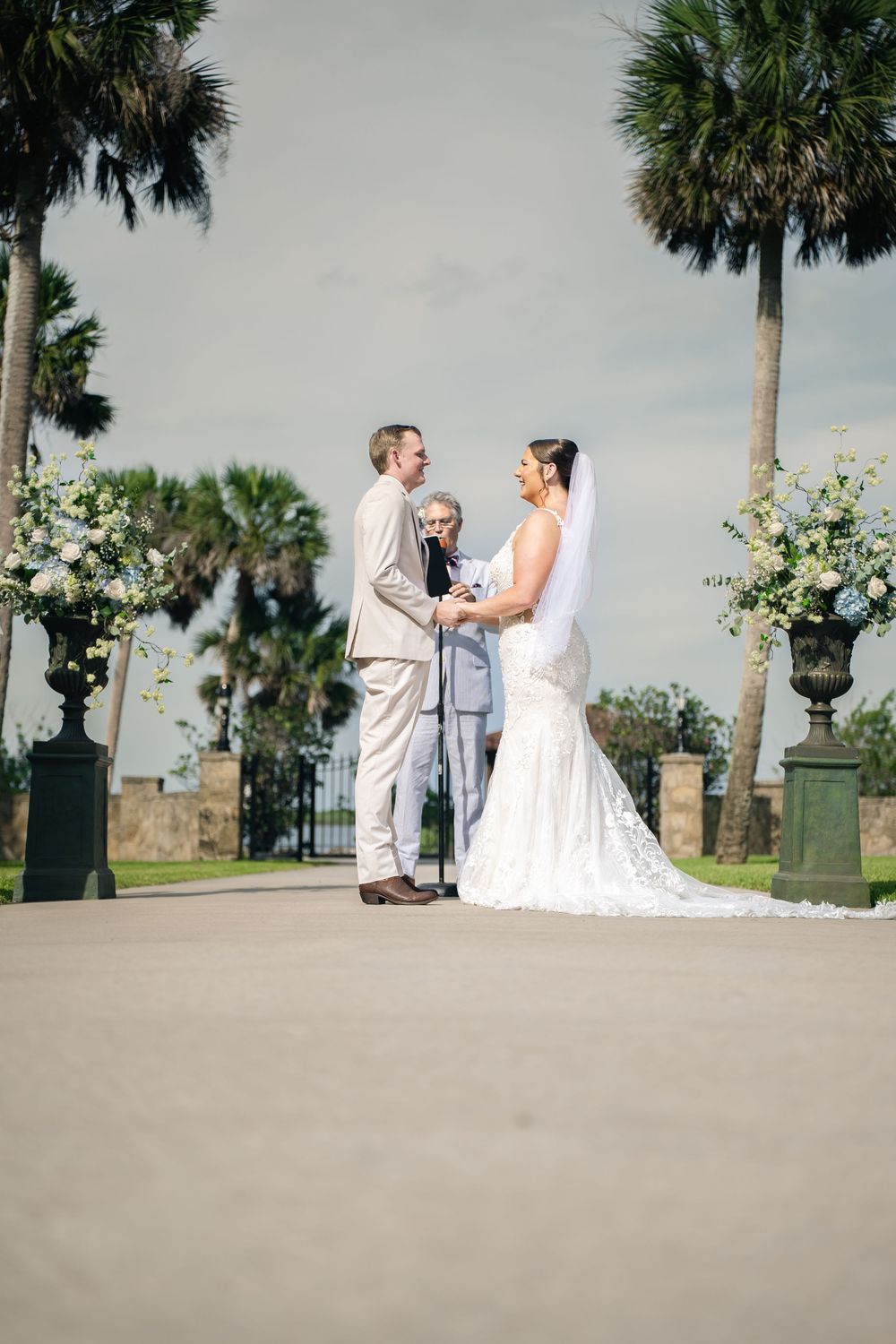 Outdoor wedding ceremony with bride in lace gown and groom in tan suit, exchanging vows before officiant amid palm trees.