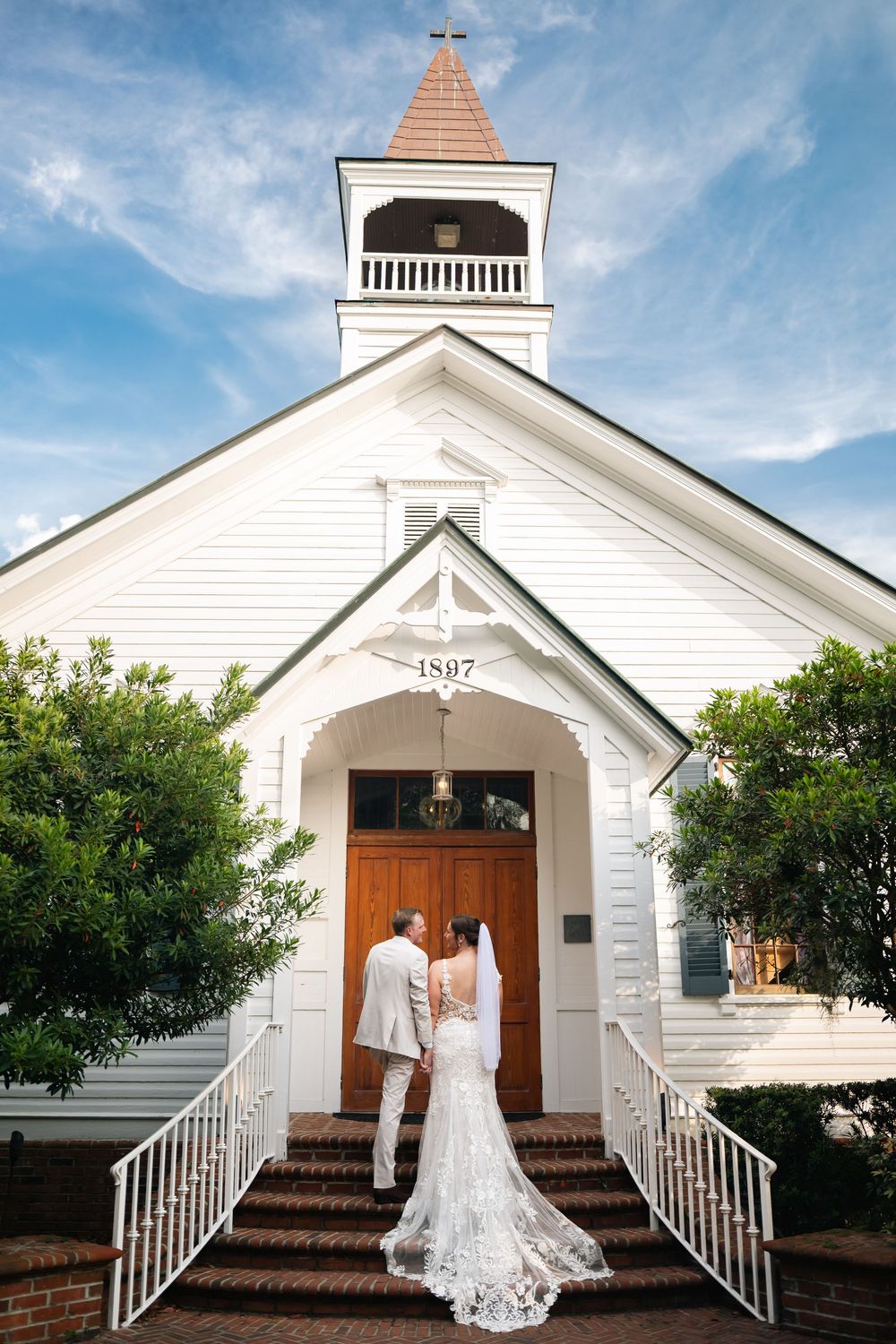 Bride and groom stand before a historic white 1897 church with a steeple on their wedding day.