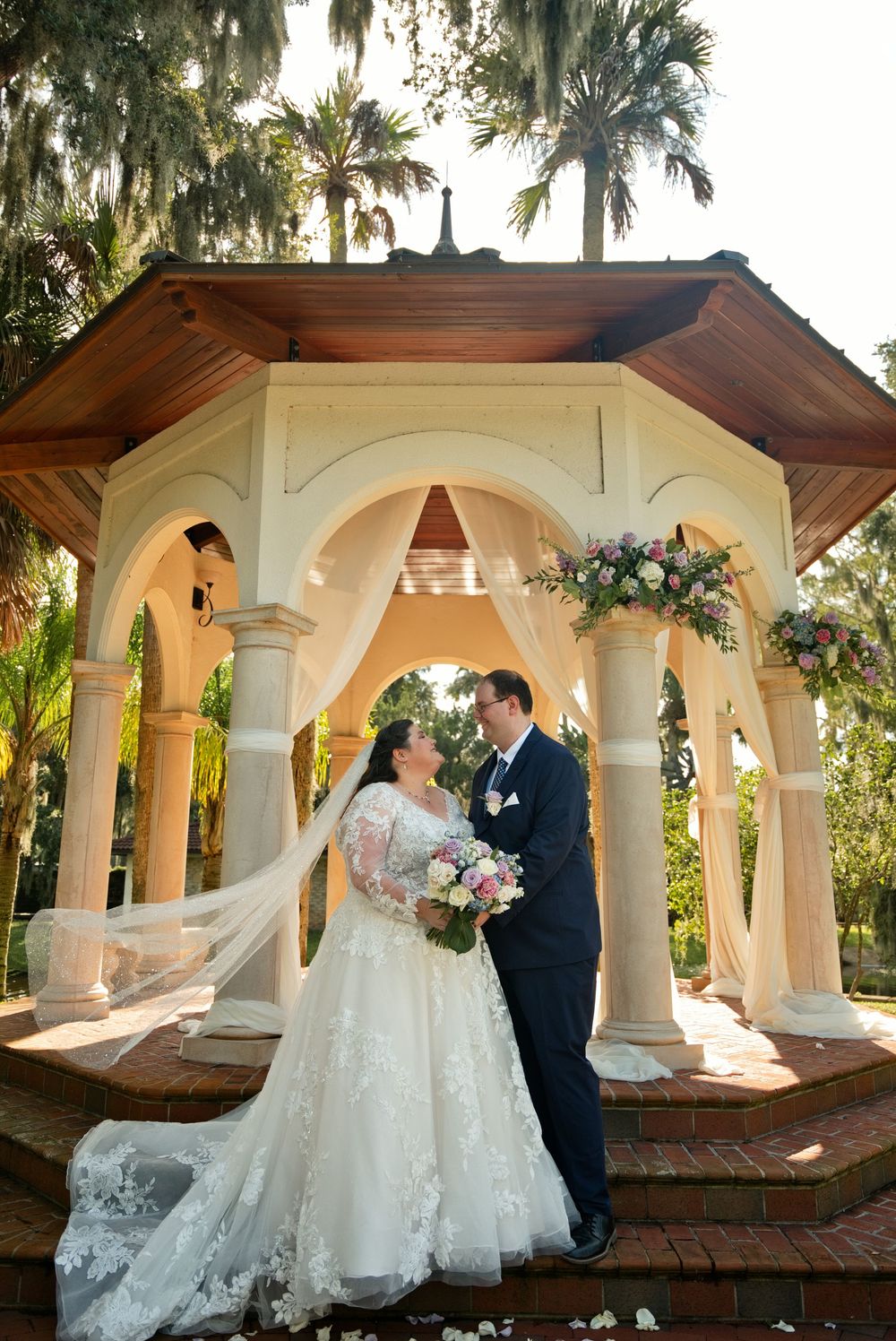 Bride and groom share a romantic moment under an elegant gazebo surrounded by lush gardens and floral arrangements.