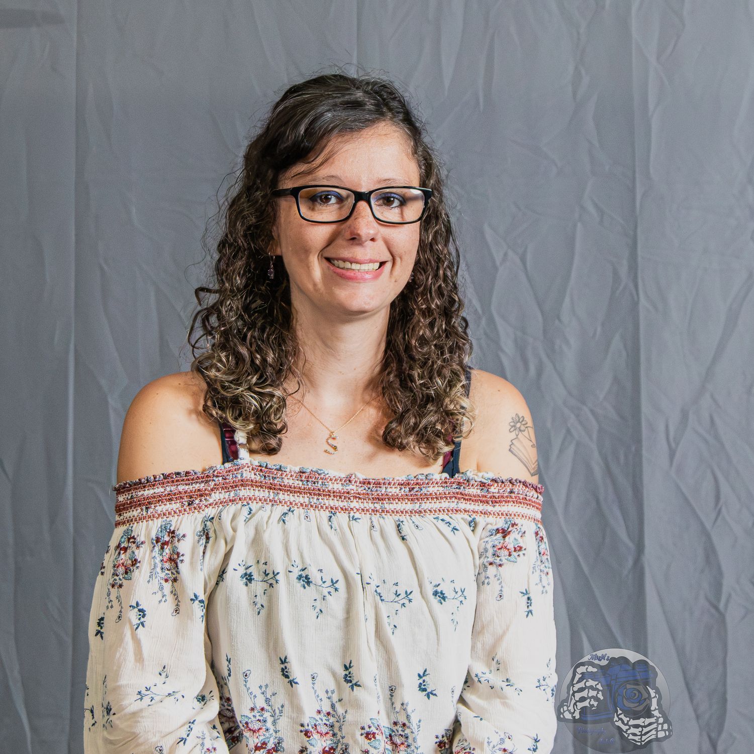 Smiling woman with curly hair and glasses wearing a floral off-shoulder top, posing against a gray background.