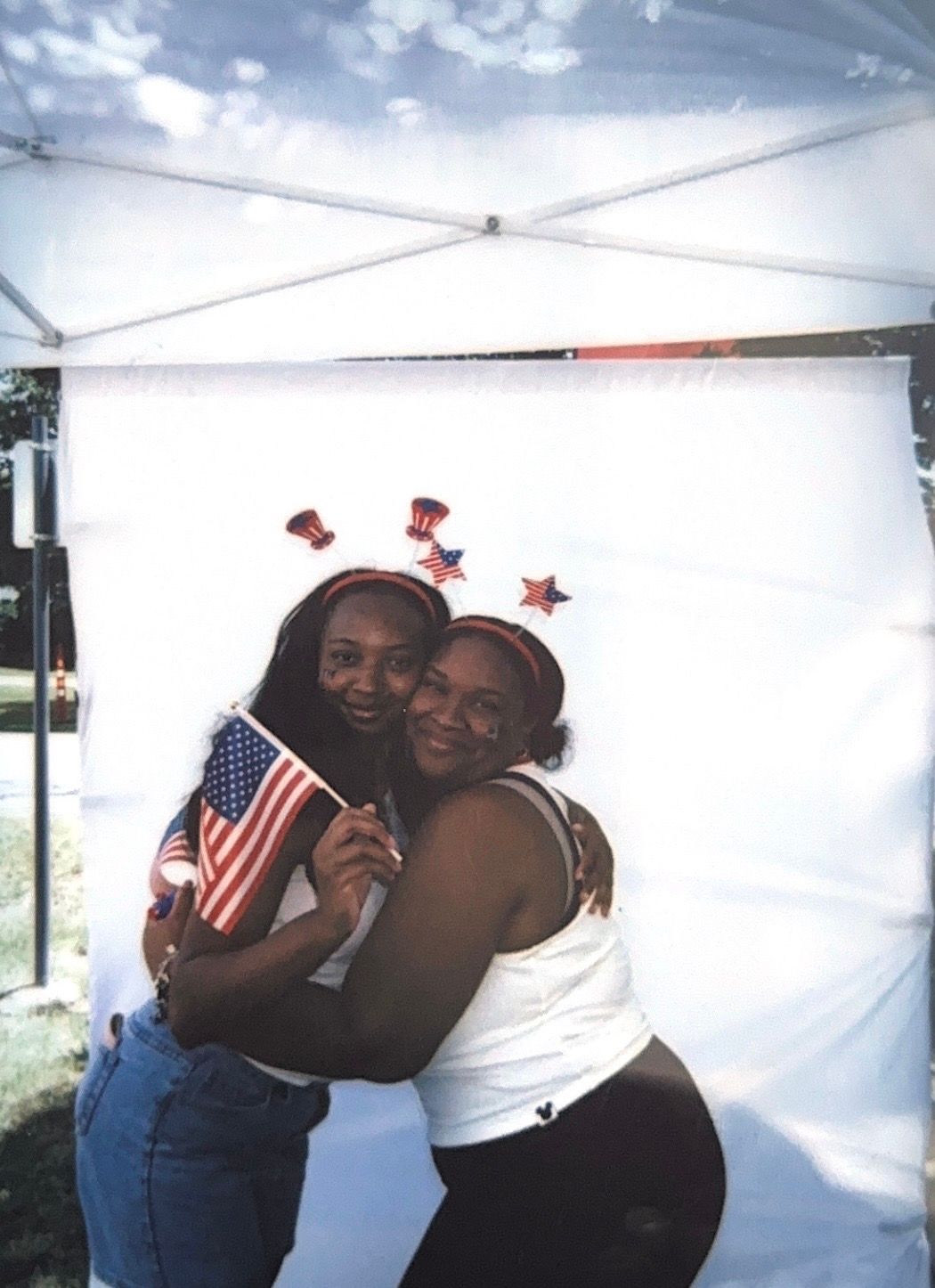 Two women hugging and smiling, holding an American flag, wearing patriotic star headbands at an outdoor Fourth of July celebration.