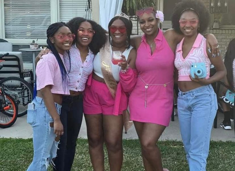 Five young women pose together outdoors, wearing coordinated pink and blue outfits, smiling at a casual backyard gathering.