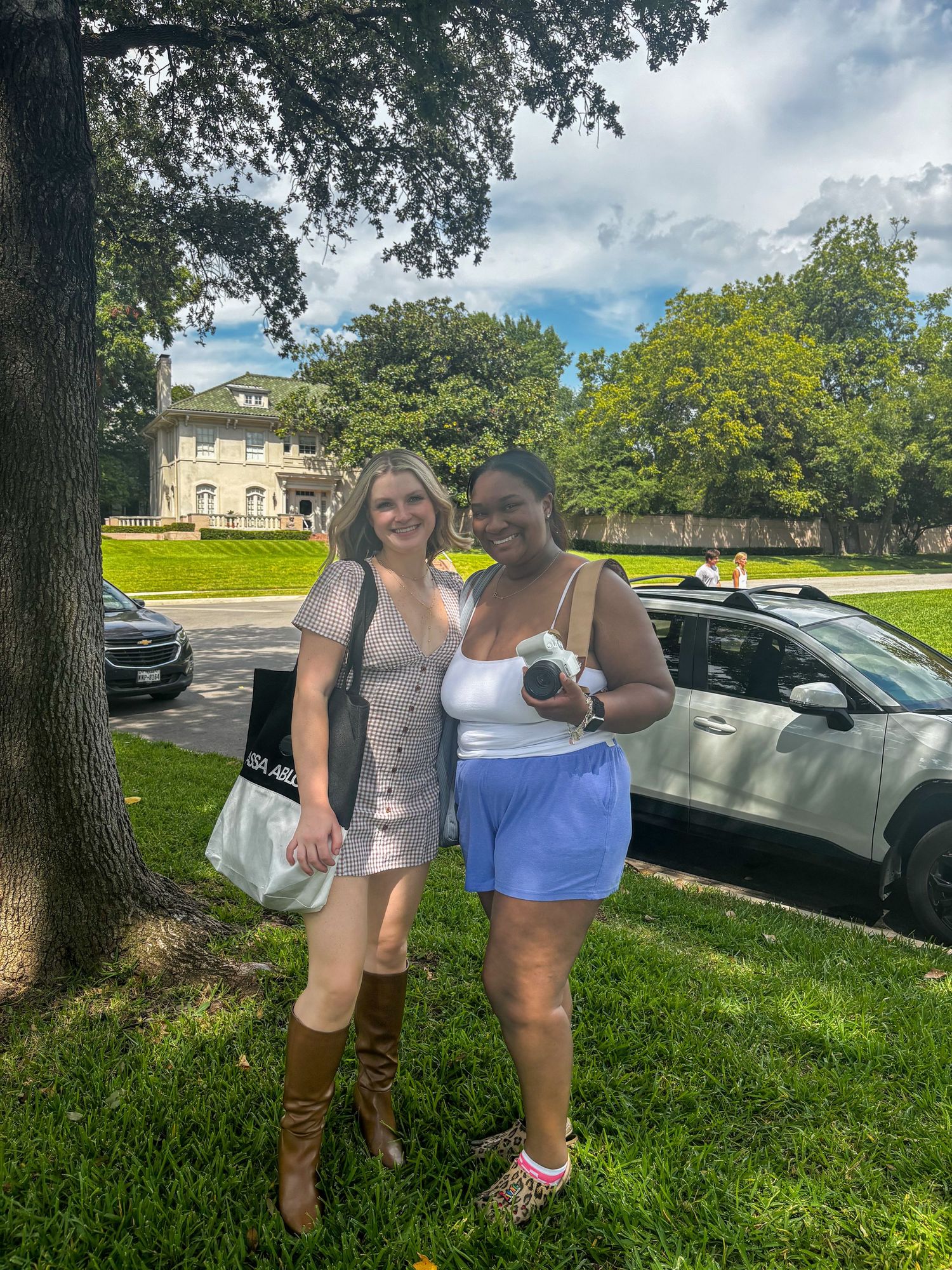 Two women smiling outdoors near trees, one in a mini dress with boots, the other in a white top and blue shorts.
