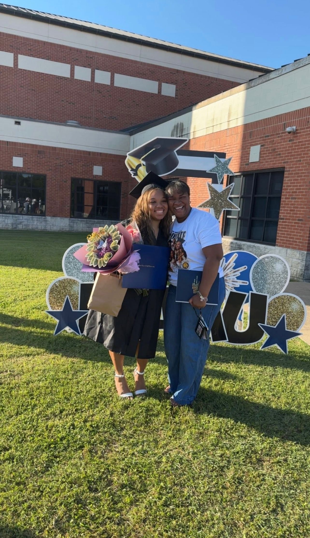 Two women pose proudly at a graduation ceremony, one in cap and gown holding flowers, in front of a brick building.