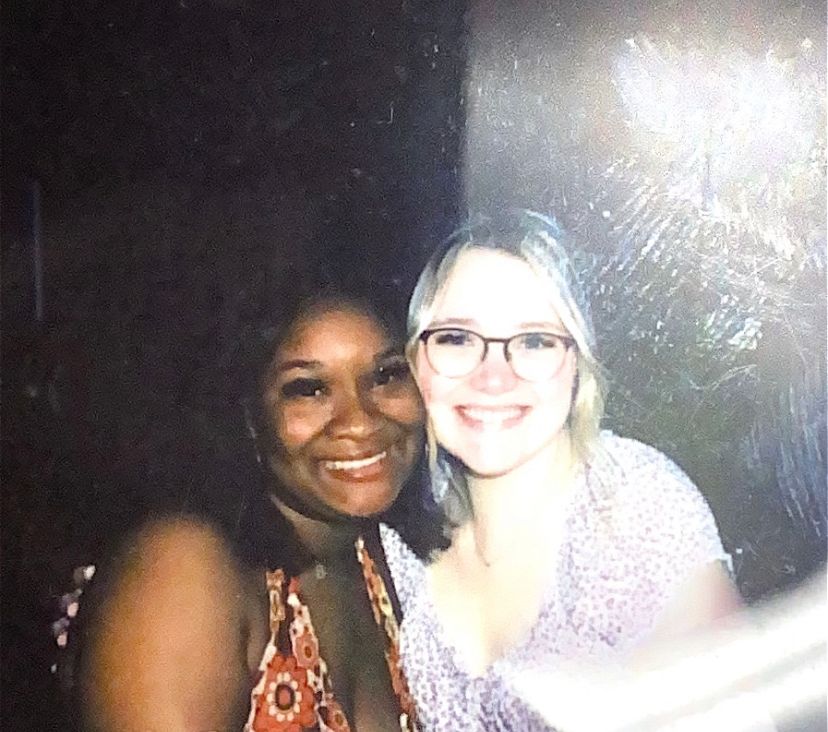 Two women smiling together for a photo at a dark indoor event, one wearing glasses and a floral top.