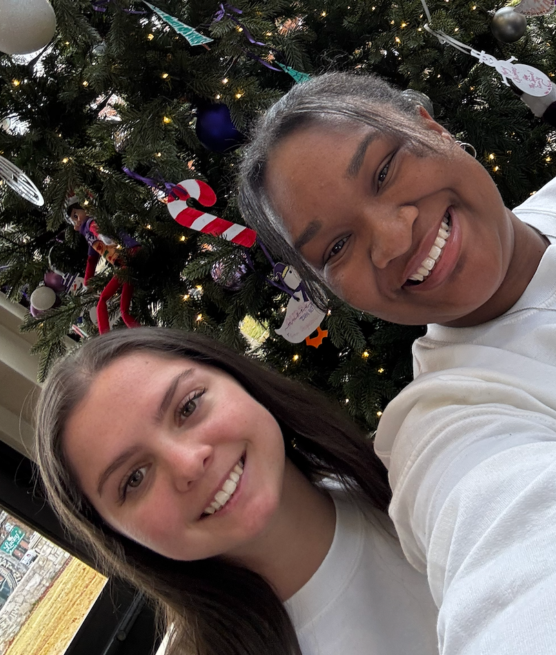 Two smiling young women take a selfie in front of a decorated Christmas tree with colorful ornaments.