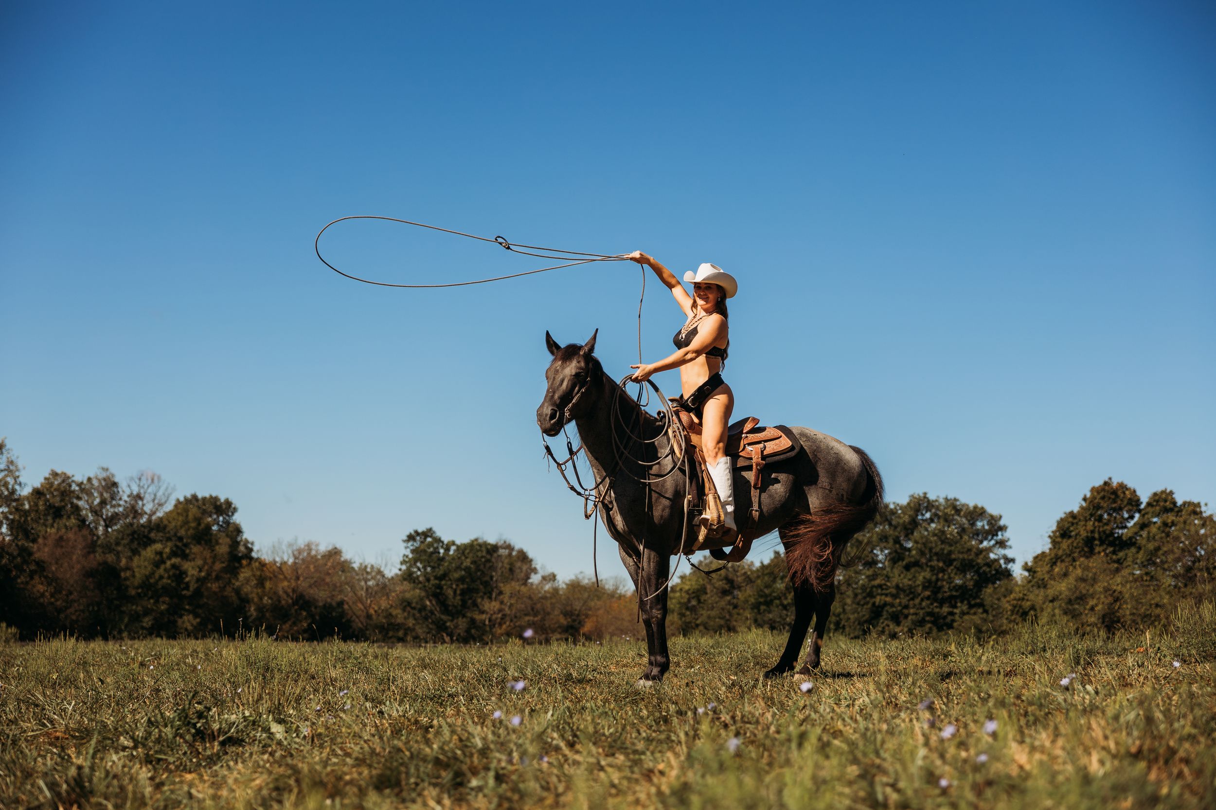 A cowgirl in a bikini and white hat swings a lasso while riding a dark horse in an open field.