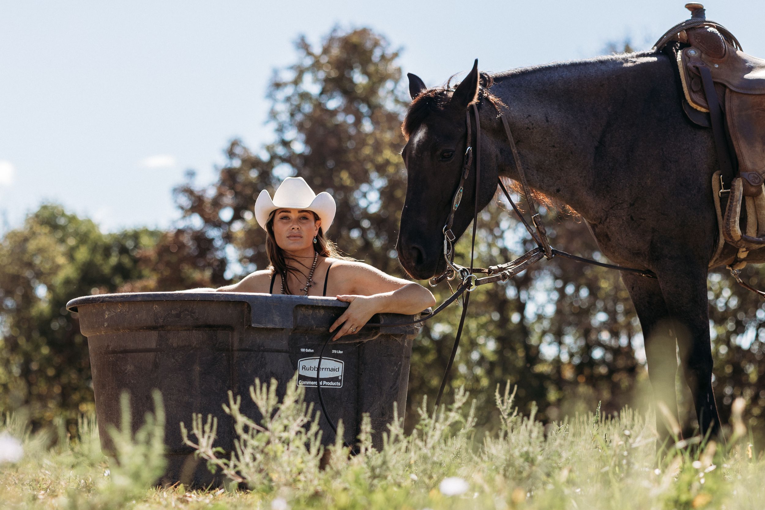 Woman in cowboy hat relaxing in outdoor tub beside a dark horse in a sunny rural field.