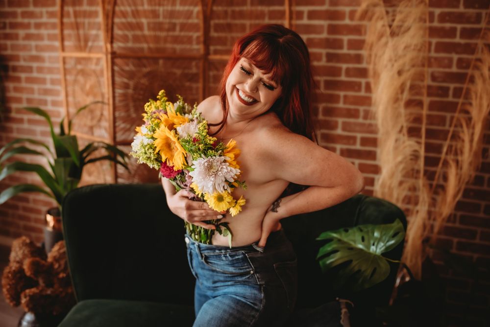 Smiling redhead woman holding colorful flower bouquet in jeans, posing in a cozy room with brick wall and green plants.