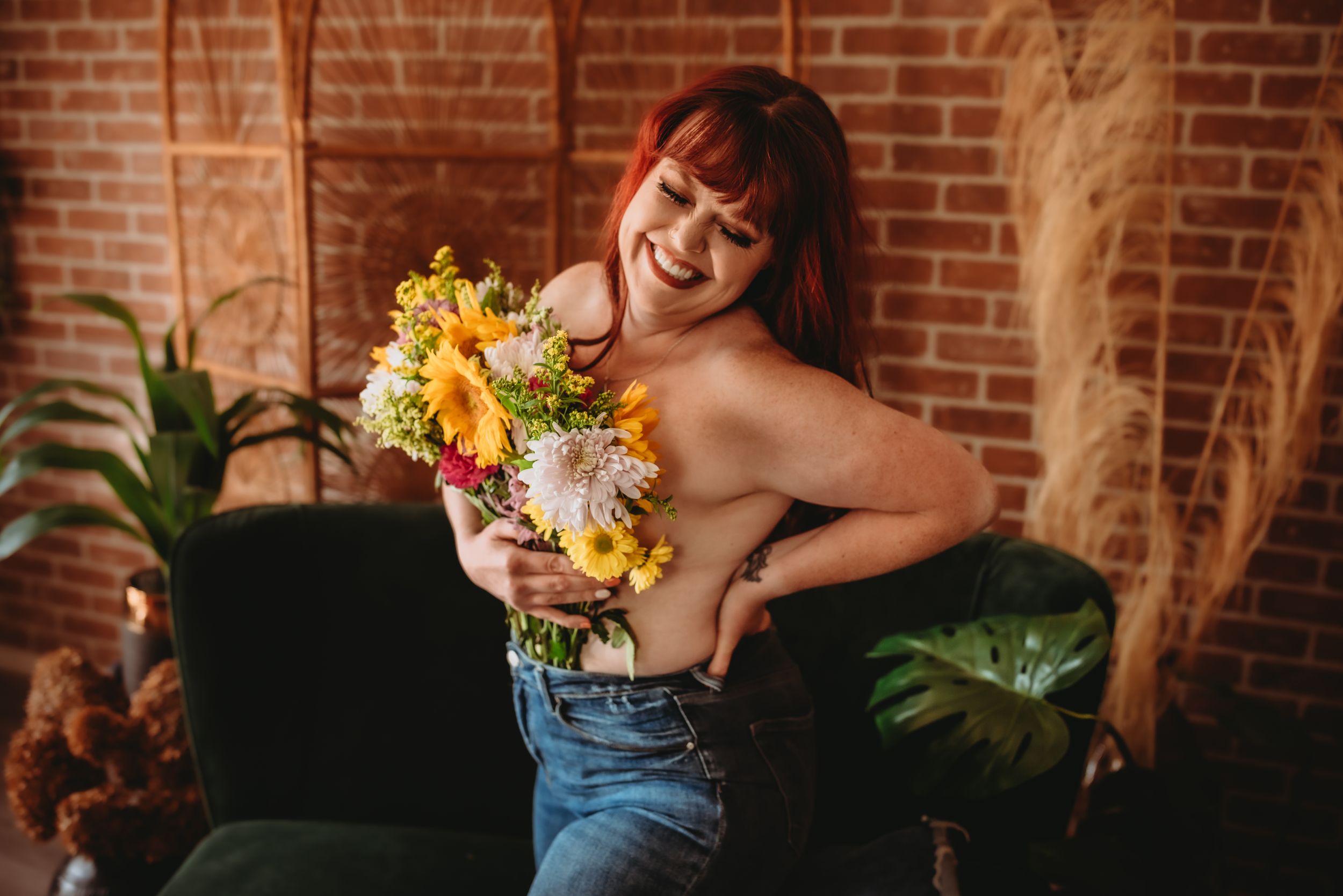 Smiling redhead woman holding colorful flower bouquet in jeans, posing in a cozy room with brick wall and green plants.