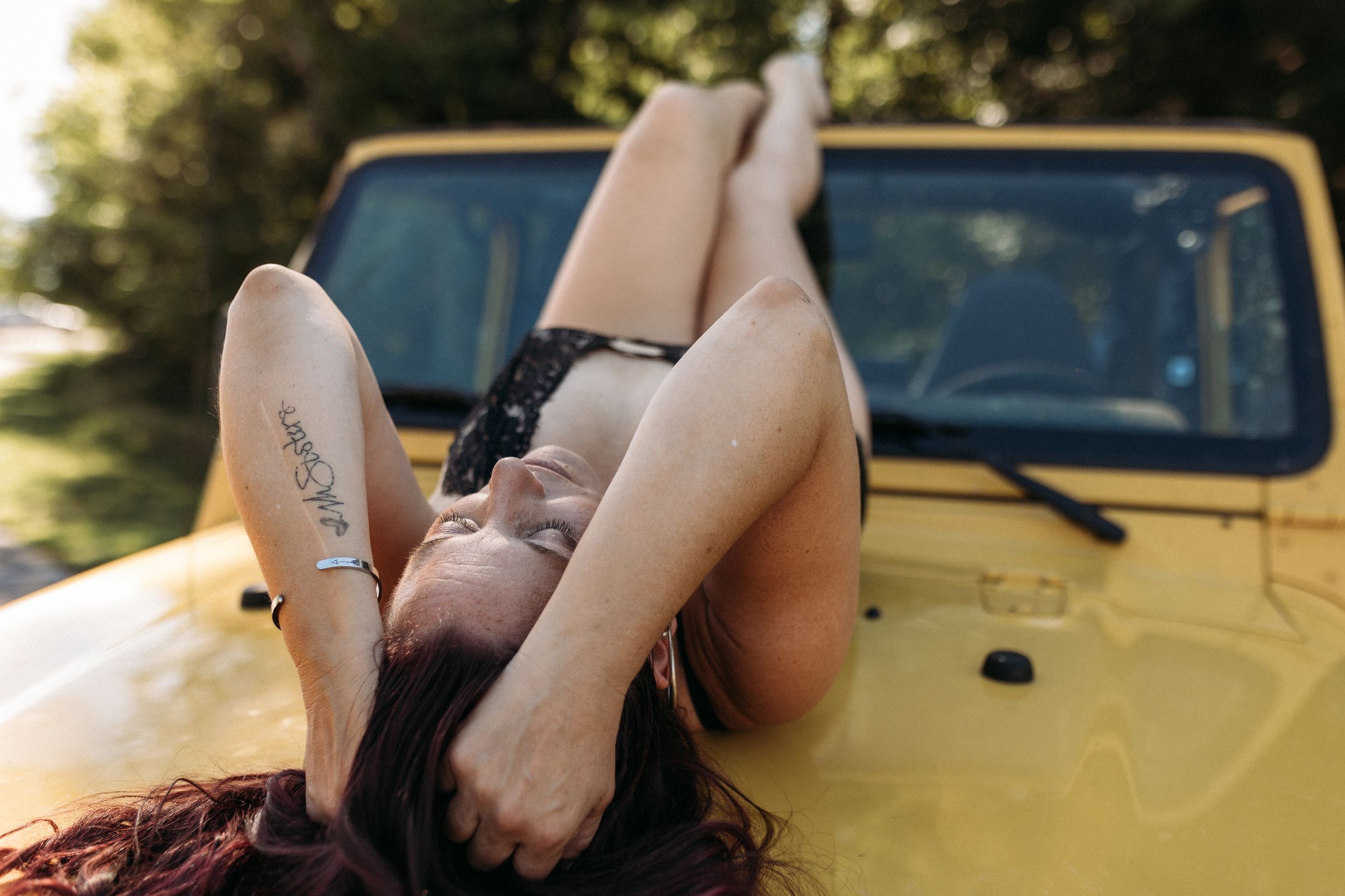 Woman with tattoo lying on yellow vehicle hood with legs raised, outdoor summer photography.