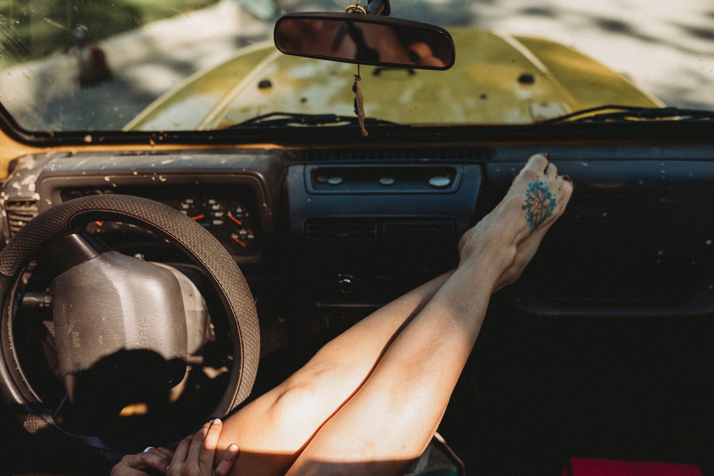 Person resting bare legs on vintage car dashboard, showing foot tattoo inside retro vehicle interior.
