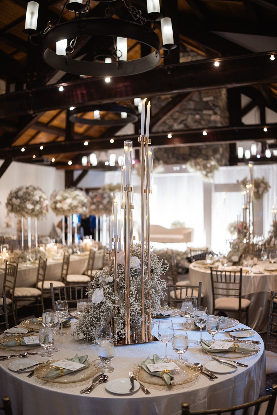 Elegant rustic wedding reception with round tables, gold candelabras, white floral centerpieces, and string lights under wood beams.