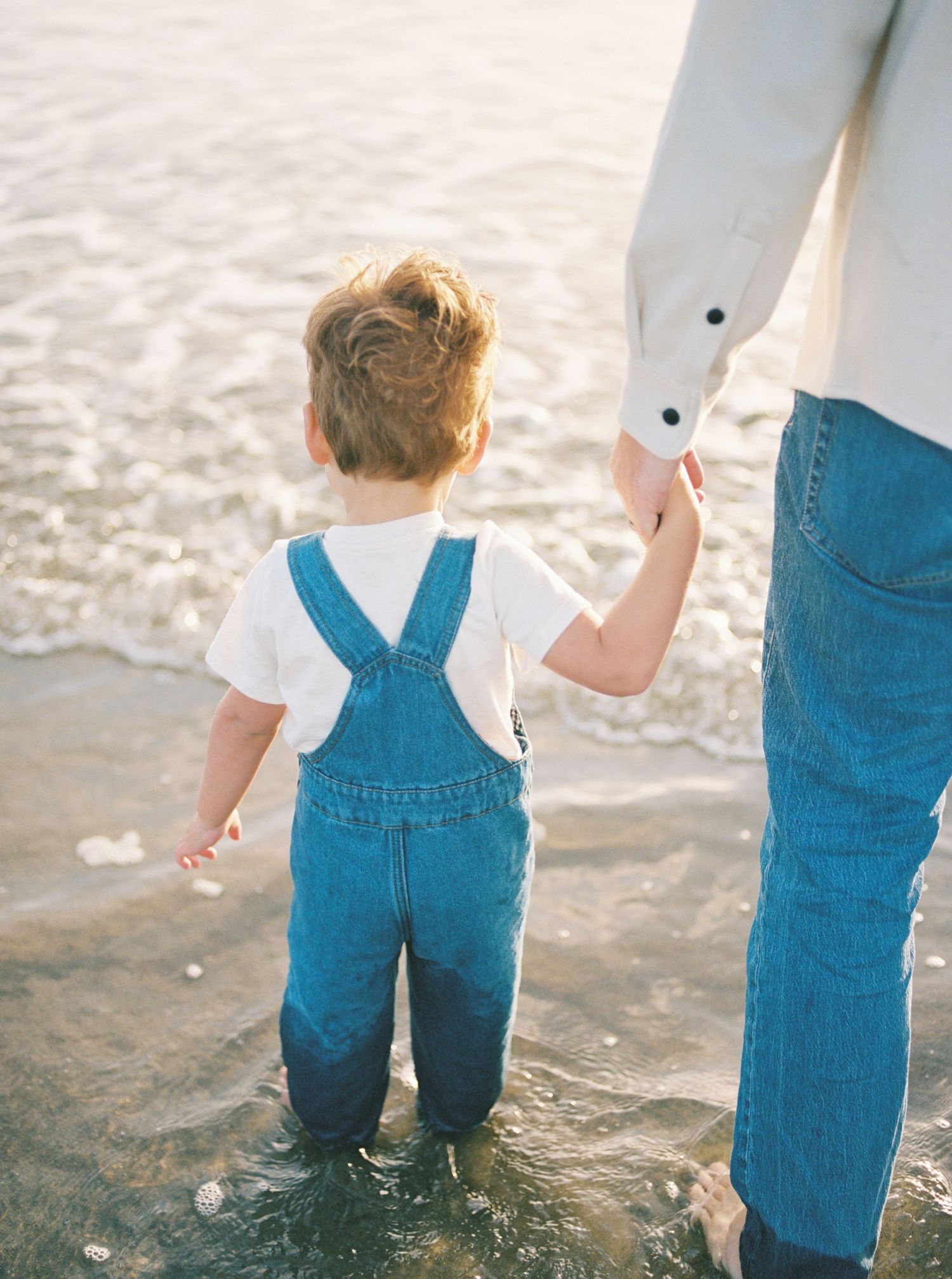 Toddler in denim overalls and white shirt holds an adult's hand while standing in shallow ocean water at the beach.