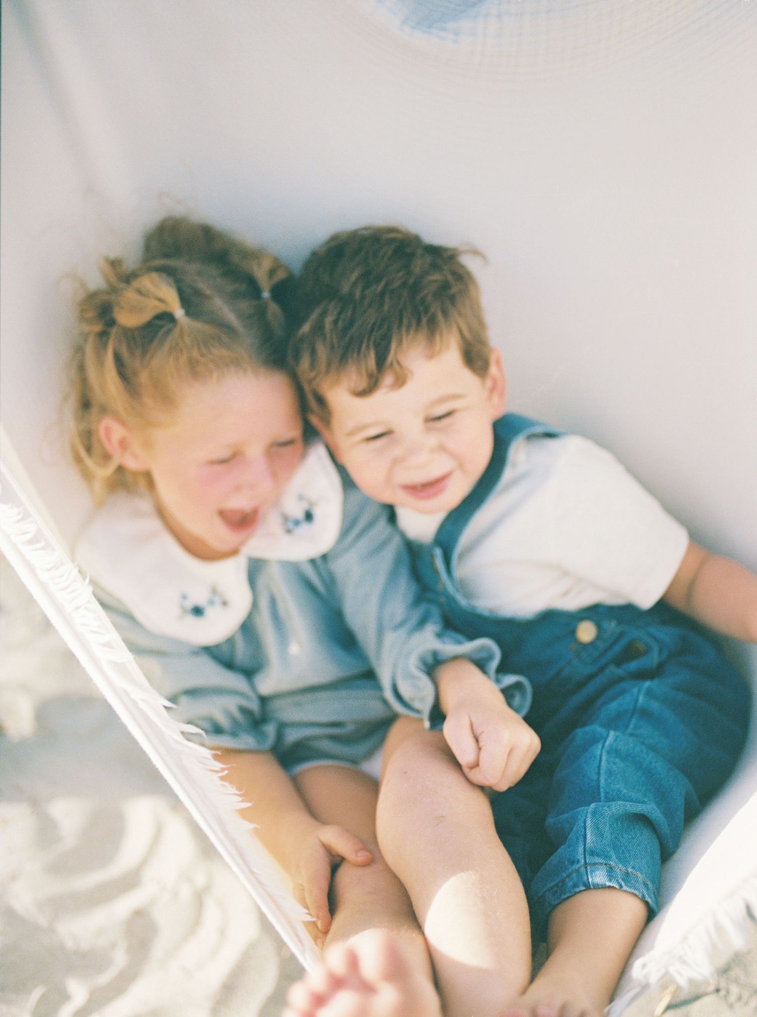 Two laughing young children in matching soft blue outfits snuggle together in a cozy hammock on a bright, sunny day.