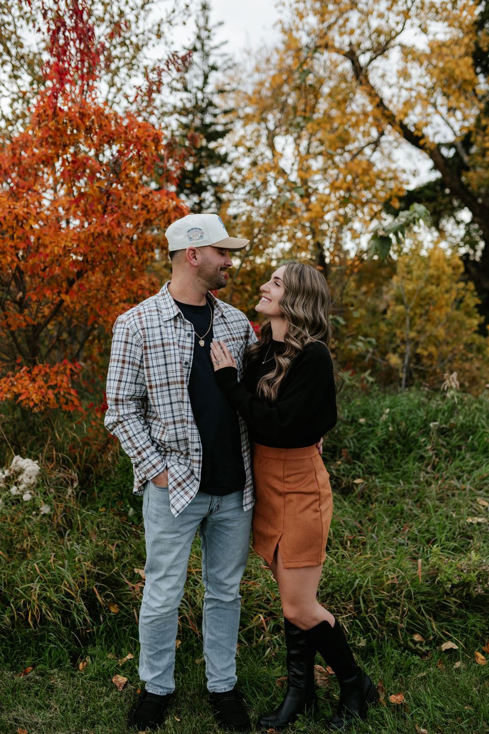 Smiling couple embracing outdoors among vibrant orange and yellow fall foliage, he in a plaid shirt and cap, she in boots and a tan skirt.