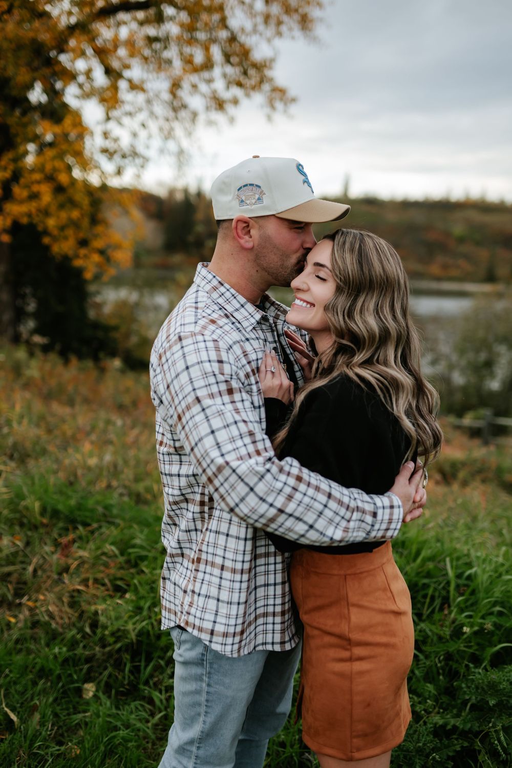 Couple embracing in a grassy field by a lake as he kisses her forehead during an autumn engagement photo session.